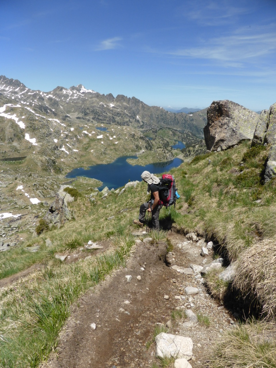 A hiker with a red backpack descending a rocky trail with a huge view over a chain of alpine lakes and snow-patched peaks