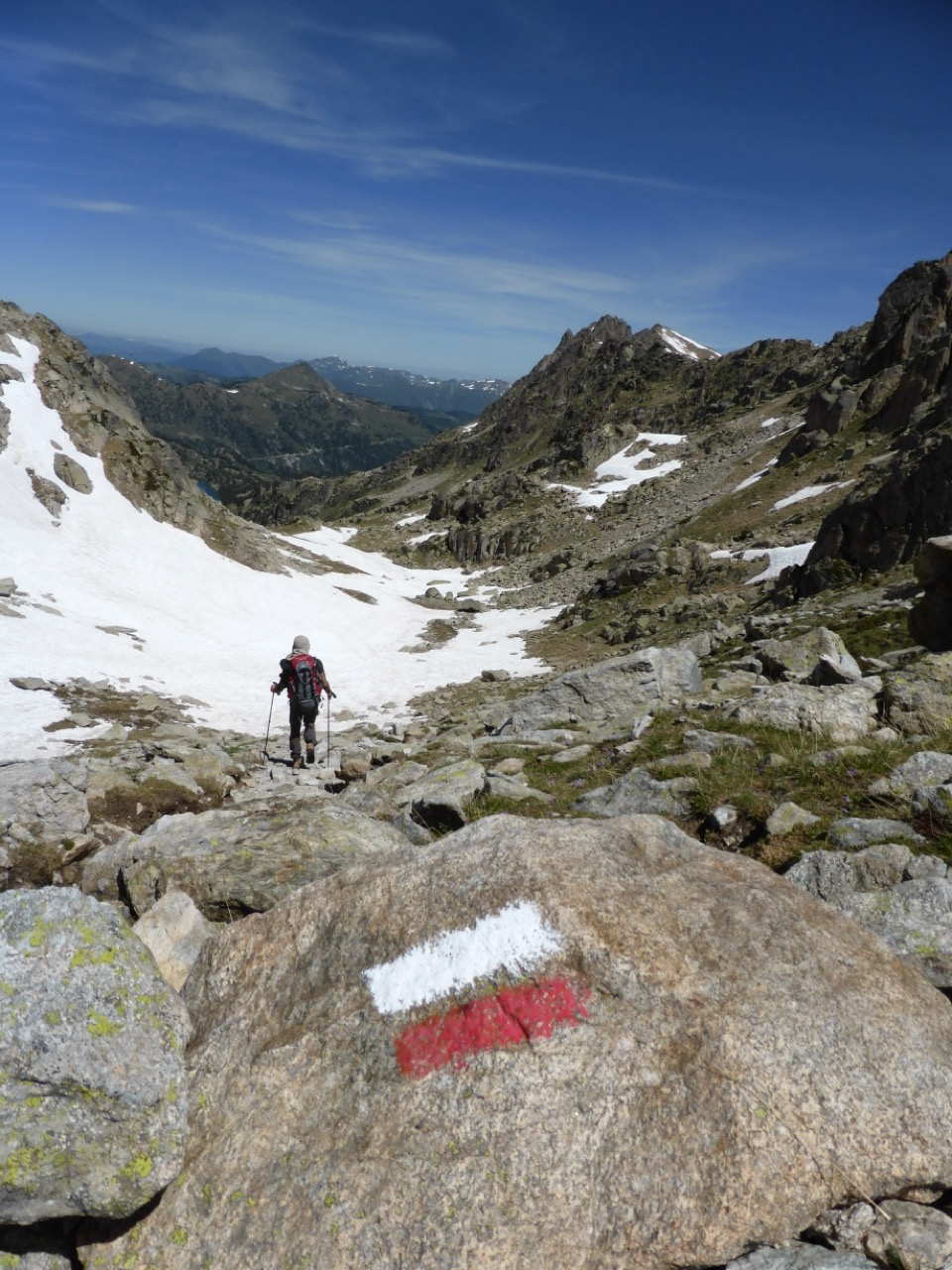 A lone hiker with a backpack descending into a rocky, snow-patched alpine valley in the Pyrenees, a red-and-white trail blaze painted on a boulder in the foreground