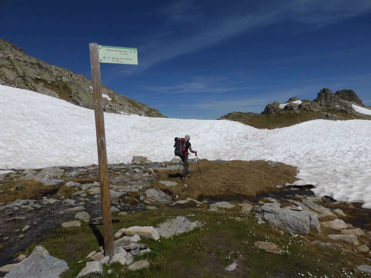 A hiker crossing a wide snow patch on a rocky mountain pass in the Pyrenees, a wooden trail sign in the foreground and a deep blue sky above