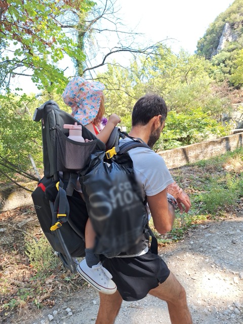 A father hiking a shaded path with a small child riding in a baby carrier on his back, wearing a floral sun hat