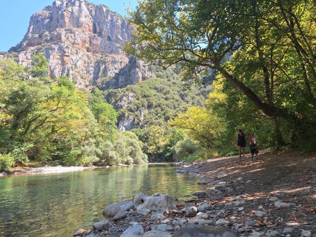 A mother and small child walking along a stony river bank under the plane-tree canopy, with a tall grey rocky peak rising above the far side of the gorge