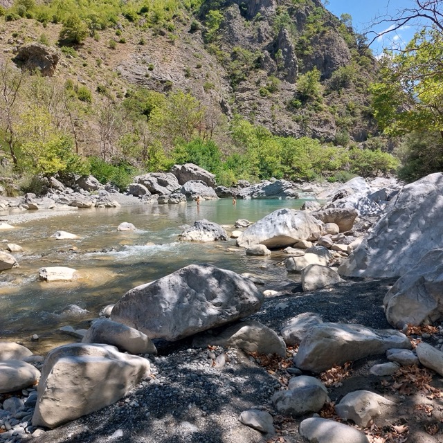 A green pool of the Aoos river at the base of a rocky gorge, with two swimmers visible in the distance and pale grey boulders piled along the near bank