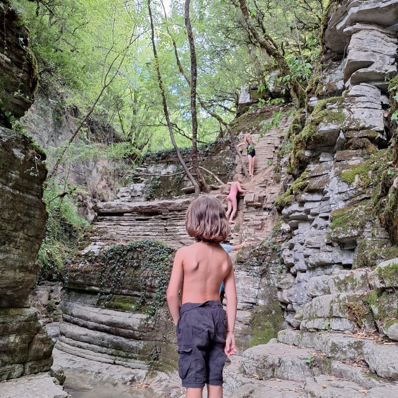 Two children climbing up the terraced limestone walls of the Kolymbithres gorge, ropes and roots for grip, with a third child standing barefoot in the shallow pool below watching them