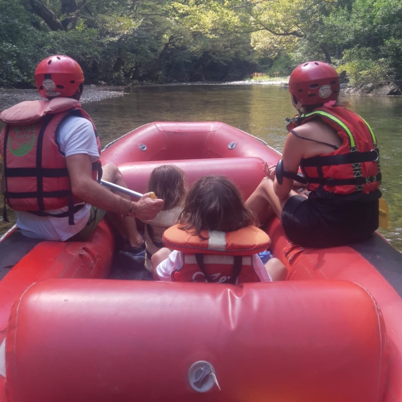 View from inside a red raft on the Voidomatis river — two adults in red helmets and life jackets paddling, two small children sitting between them, calm turquoise water and plane-tree canopy ahead