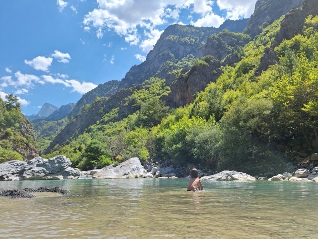 A swimmer floating in a clear green pool of the Aoos surrounded by steep forested slopes, with dramatic limestone peaks rising above the tree line under a blue sky with scattered clouds