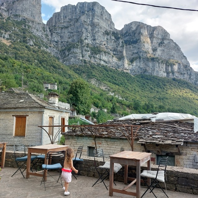 A small child walking across a stone-paved terrace in Papingo, with the sheer limestone wall of the Astraka Towers rising behind the village