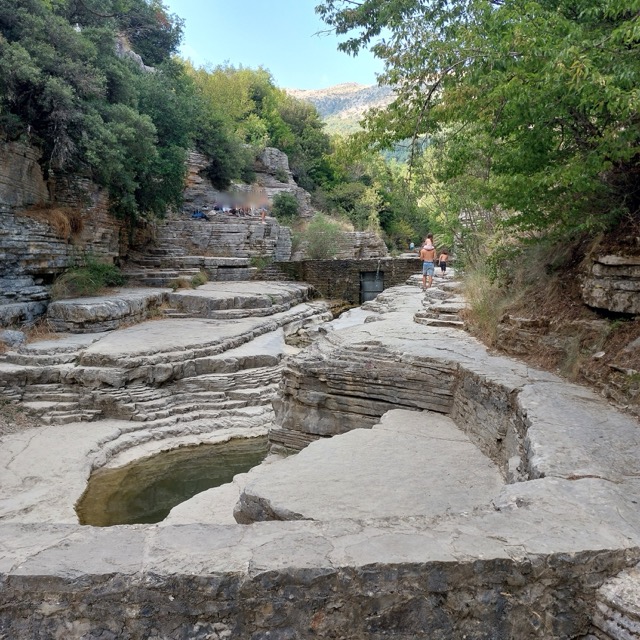 A shallow gorge with a stone pathway winding past a series of natural pools cut into terraced grey limestone, with small groups of visitors in the distance