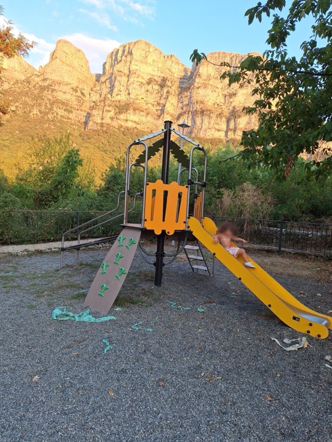 A child on a yellow slide in a small mountain playground, with the grey limestone walls of the Astraka Towers rising in the background at sunset
